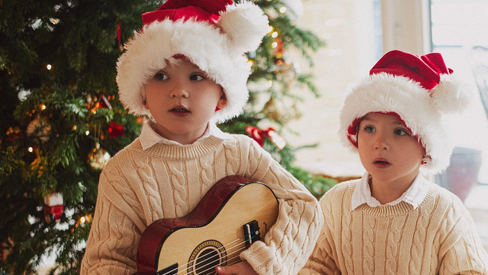 To børn i julemandshuer, hvor den ene spiller guitar, sidder ved juletreet.