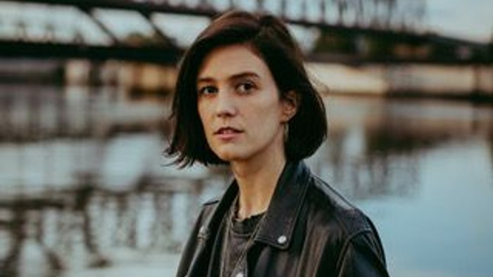 Eine Frau in einer Lederjacke steht in der Nähe eines Gewässers mit einer Brücke im Hintergrund.