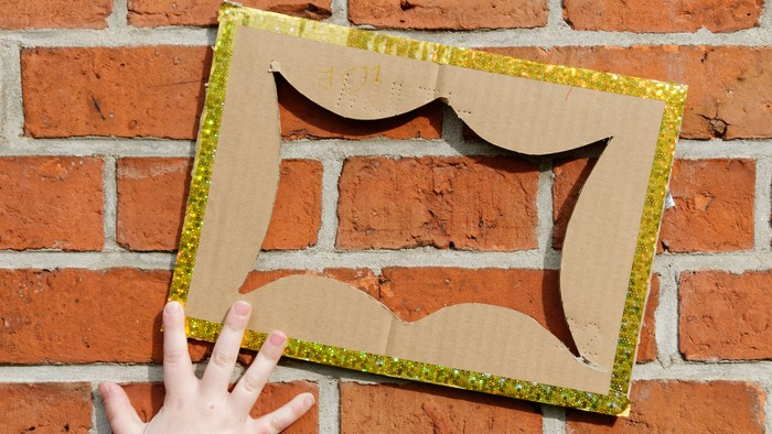 Eine Hand hält einen selbstgemachten Bilderrahmen aus Pappe und Glitzerfolie an einer Ziegelwand