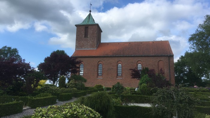 En rød murstenskirke med tårn og grøn spire, omgivet af en have med buske og blomster.