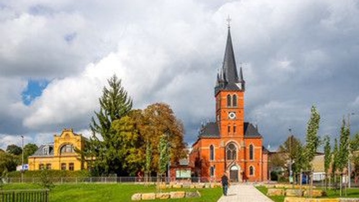 Eine malerische Kirche mit hohem Turm erhebt sich inmitten eines ruhigen, gestalteten Stadtplatzes unter dramatischem Himmel.