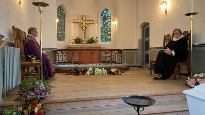 En lille kirke med to personer, en præst og en kvinde, sidder på bænke. Alteret med kors og blomster.