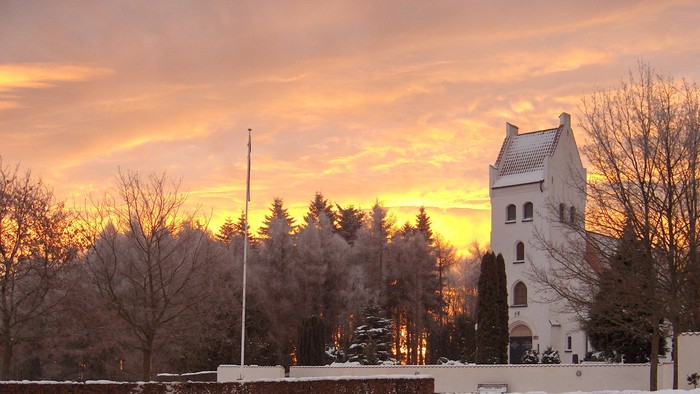 En kirke med tårn og en flagstang foran, omgivet af snebede træer og en rødgul solnedgangshimmel