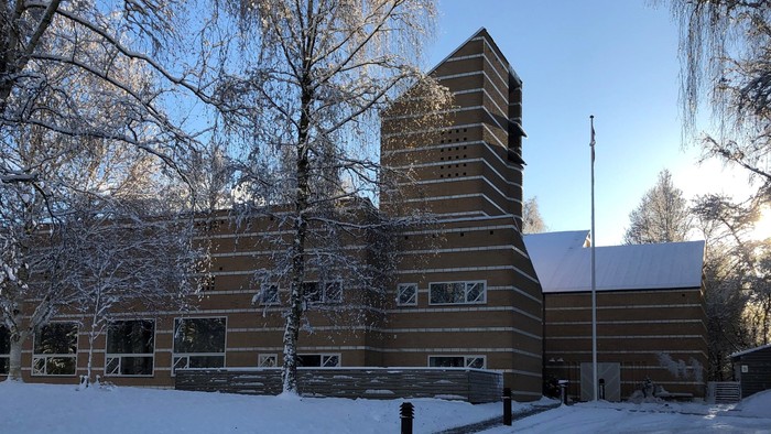 En snedækket vej forbi et stort, mørkt træhus med tårn og bjærketræer.