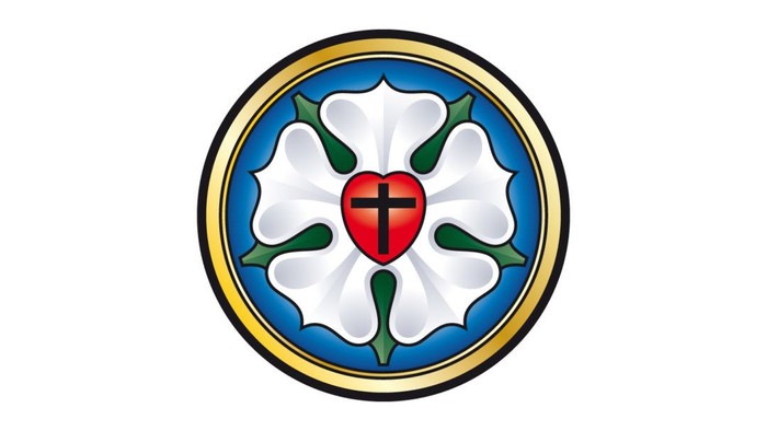 Das Bild zeigt ein Emblem mit einem roten Herz und einem Kreuz auf weißem Hintergrund, umgeben von grünen Blättern und einem blauen Ring.