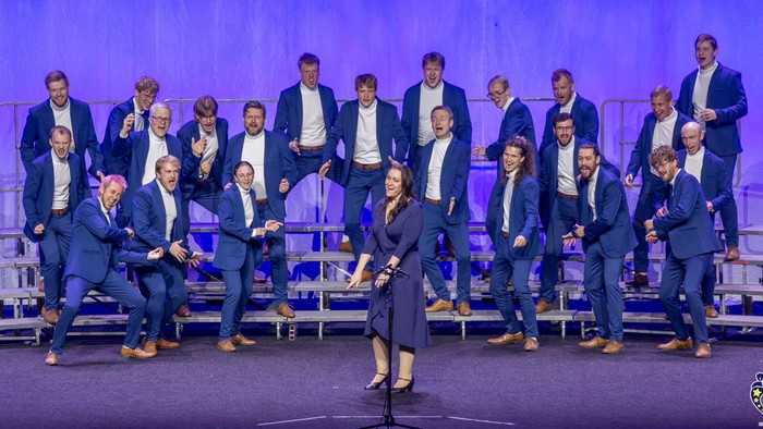 Gruppe af mænd i jakkesæt, der optræder på scenen, med en kvinde, der synger forrest.