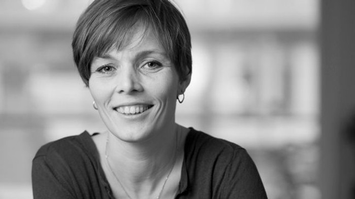 Selvsikker professionel kvinde, der smiler med armene krydset, siddende ved et bord i et lyst indendørs miljø.