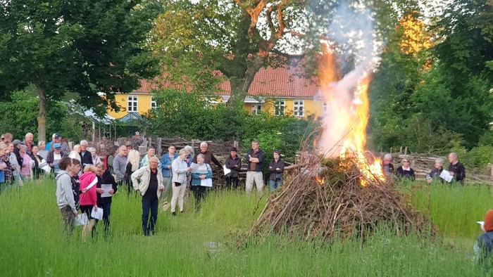 En gruppe samles omkring et brændende bål på en græsklædt udendørsplads med huse i baggrunden.
