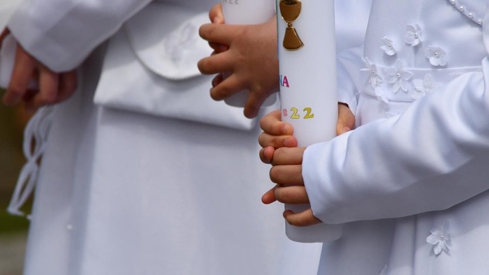 Kinder in weißen Kommunionkleidern halten verzierte Kommunionkerzen in den Händen