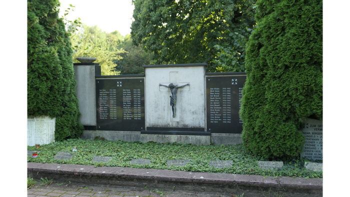 Ein Kreuz mit einer Figur in einem grünen, gepflegten Bereich mit einer Mauer und Bäumen.