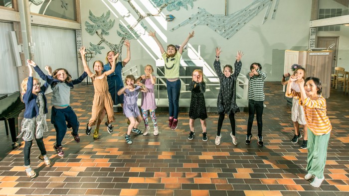 En gruppe unge danser og springer glade i en hall med mosaikvæg.