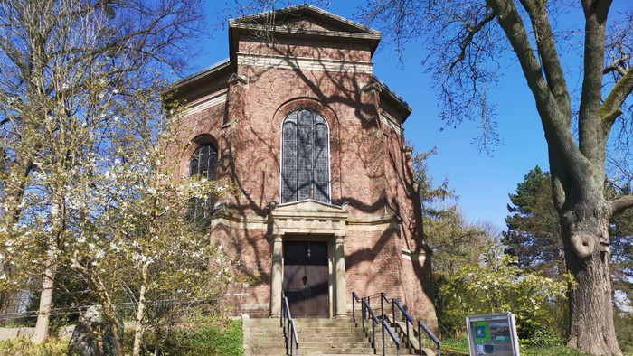 Das Bild zeigt eine alte Steinstruktur mit Treppe und Fenster, umgeben von Bäumen und Büschen.