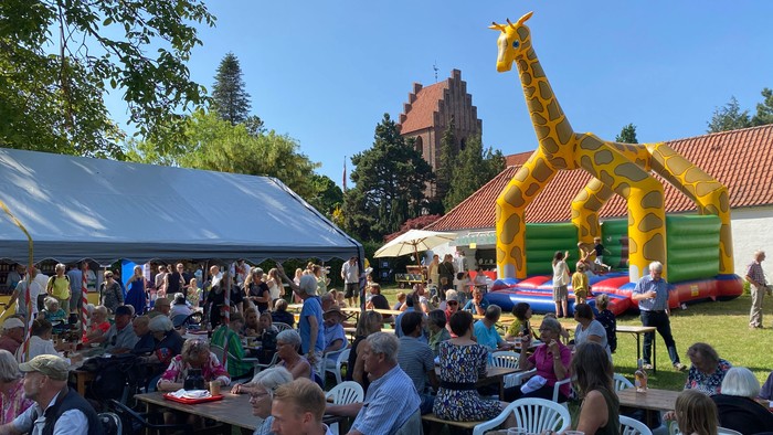 En folkelig fest med mange mennesker, der sidder ved bord og spiser, og en stor opblæst giraf i baggrunden.