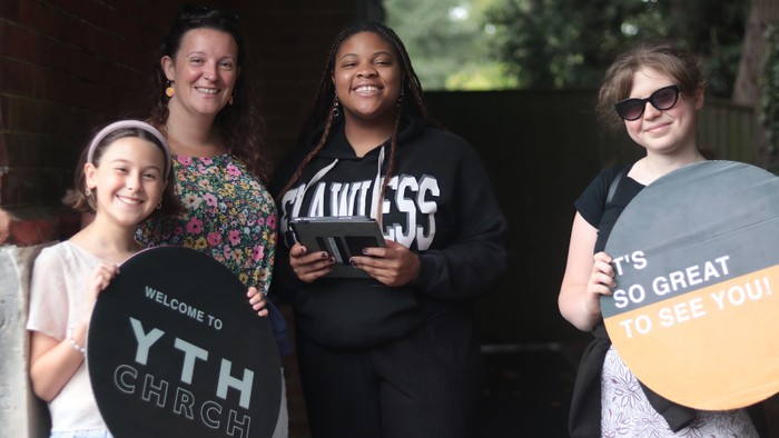 Four women pose together, two holding signs welcoming visitors to YTH Church.