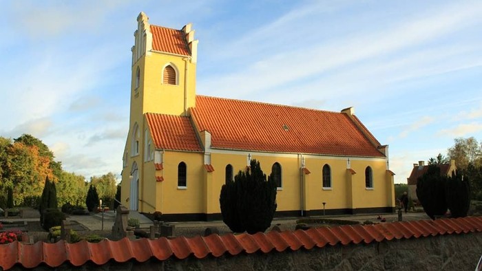Gul kirke med rødt tag og tårn, omgivet af træer og mur.