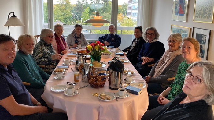 En gruppe af ældre personer sidder til middag i et lysligt og rummeligt stue.