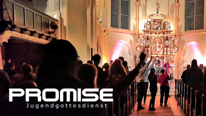 Eine Gruppe von Menschen singt in einer Kirche, wobei ein großes, verziertes Kreuz im Hintergrund steht.