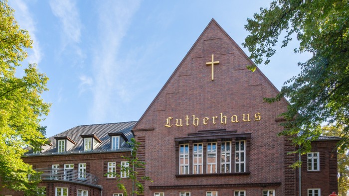 Das Bild zeigt ein großes, rotes Gebäude mit einem Kreuz und der Aufschrift 