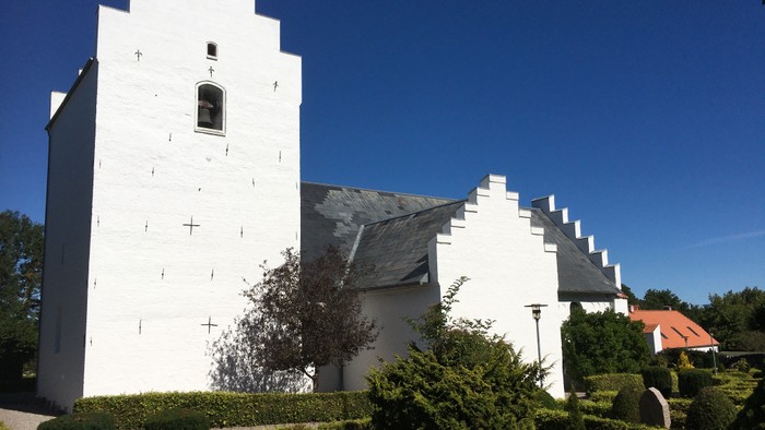 Hvid kirke med tårn og spiret, omgivet af grønne buske og et blåt himmel.
