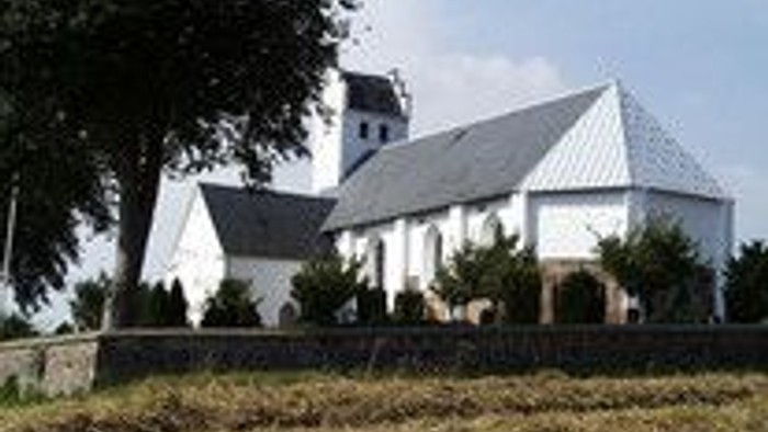 En lille hvid kirke med tårn og en mindre bygning ved siden af, omgivet af en hegn og træer.