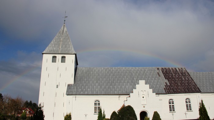 Nørre Løgum kirke, set fra syd med blå himmel og gråhvide skyer og en regnbue som baggrund. Kirken er hvidkalket, har et tårn og der er bly på taget. Forrest ses grønnehække og buske på kirkegården