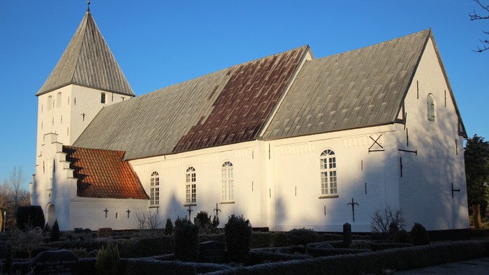 Kirken set fra syd-øst med frostblå himmel som baggrund. Der er skygger af træerne på kirkens hvidkalkede facade, og på de små hække ses frost på toppen.
