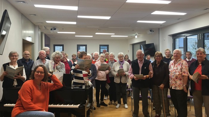 Eine Gruppe älterer Menschen singt gemeinsam in einem Raum, begleitet von einer Frau am Klavier.