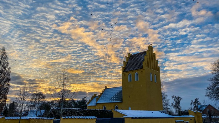 Gul kirke med tårn og spiret under snødekt himmel med solopgang