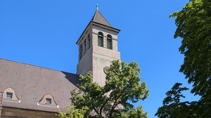 Kirchturm mit Uhr und Kreuz, umgeben von Bäumen und Gebäuden