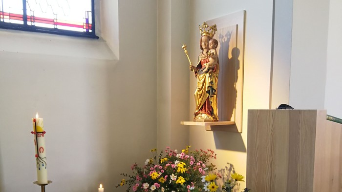Auf einem Altar stehen eine Statue, Blumen und Kerzen vor einem Fenster