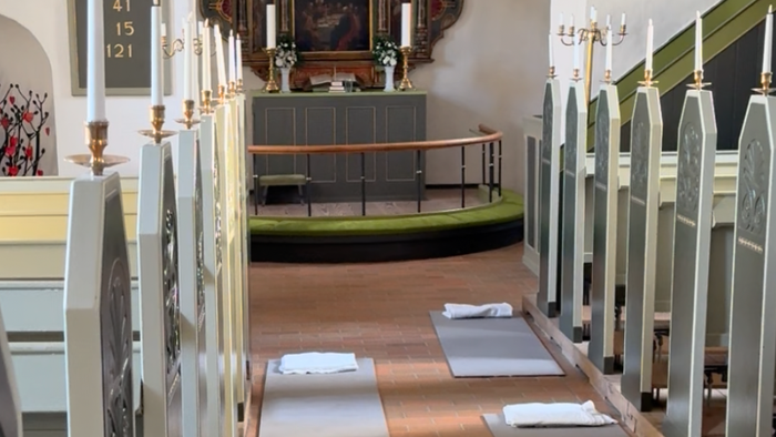 Et roligt kirkeinteriør med træbænke, hver dækket med hvide puder, der fører hen til alterområdet.