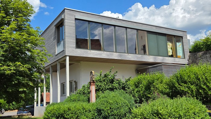 Modernes zweistöckiges Haus mit großen Glasfenstern, auf Pfeilern erhöht, umgeben von üppigem Grün.