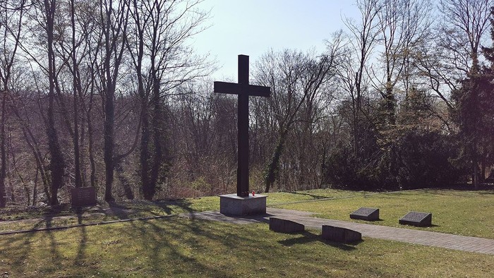 Ein hohes Kreuz steht auf einem grasbewachsenen Hügel mit Bäumen im Hintergrund.