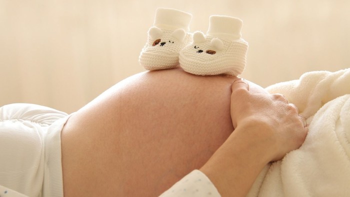 Schwangere Person liegt mit Händen auf dem Bauch, darauf liegen kleine gestrickte Babyschuhe mit Tiergesicht.