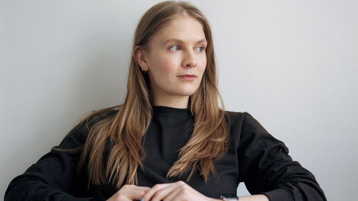 En person med langt hår sidder eftertænksomt i en sort top foran en ensfarvet baggrund.