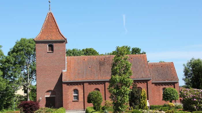 Røde murstenskirke med tårn og spiret, omgivet af grønne træer og blomster.