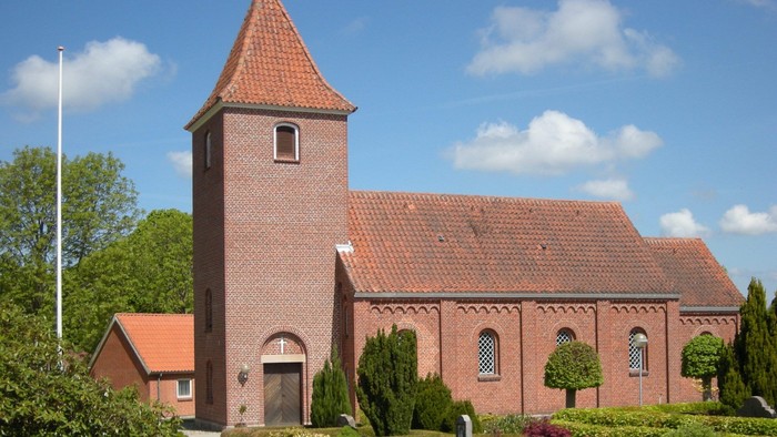 En rød murstenskirke med tårn og omgivet af grønne planter og blomster.
