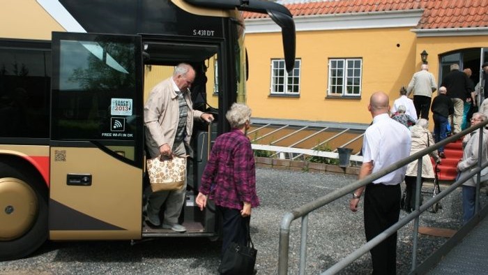En gruppe ældre mennesker går af en bus ved et gult hus