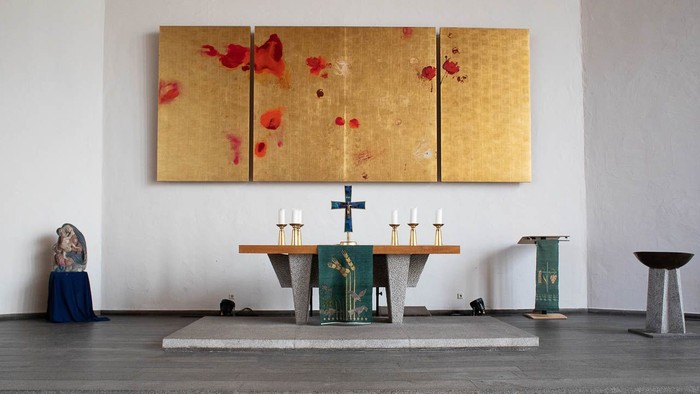 Ein Kircheninneres mit Altar, Kerzen und einem großen goldenen Bild mit roten Blumen an der Wand