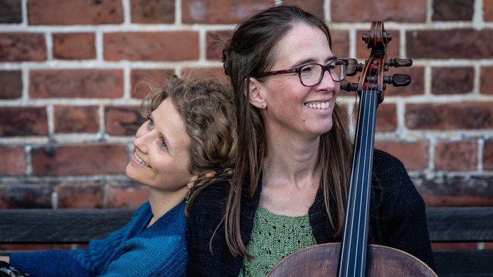 To kvinder smiler, den ene holder en cello op ad en murstensvæg.