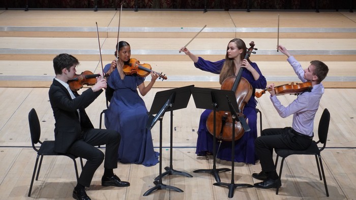 String quartet performing on stage with violins, cello, and conductor-like gestures.