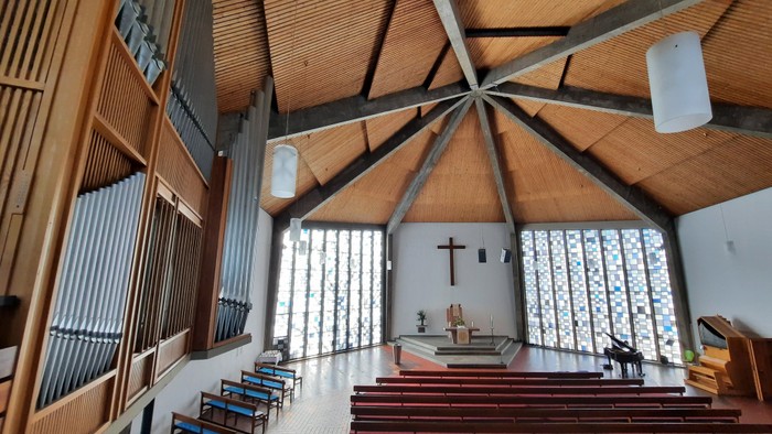 Moderne Kircheninnenraum mit hölzernen Kirchenbänken, Kreuz und großen Fenstern.