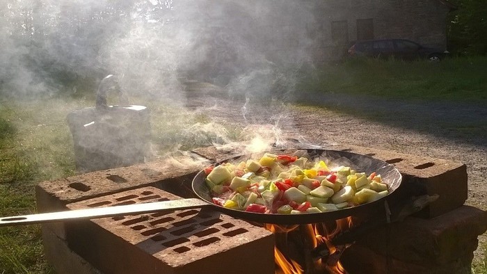 Auf einem Holzkohlegrill wird Gemüse in einer Pfanne gebraten, umgeben von Rauch und Flammen.