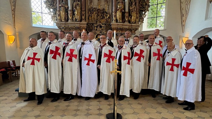 Gruppe af mænd i hvide kjortler med røde kors, stående i en kirke til en formel sammenkomst.