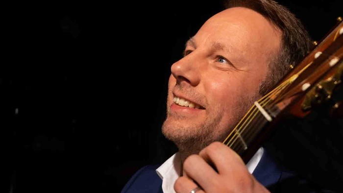 Ein lächelnder Mann spielt eine akustische Gitarre in einer dunklen Umgebung, gekleidet in einen Anzug und ein weißes Hemd.