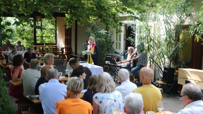 Eine Gruppe von Menschen hört einem Mann mit Gitarre und einer Frau mit Mikrofon zu, die auf einer Bühne unter freiem Himmel spielen.
