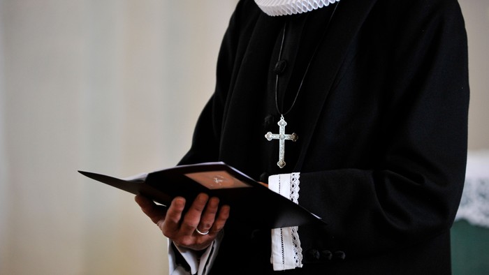 En person i sort kåbe holder en åben bog og bærer et korshalskæde.