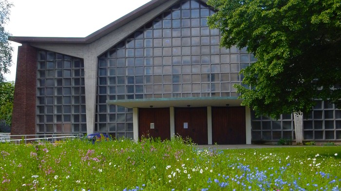 Moderne Kirche mit Glasfassade und Kreuz auf dem Dach, umgeben von blühendem Gras und Bäumen