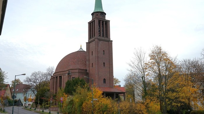 Kirchturm mit grünem Dach vor rotem Backsteingebäude mit rundem Kuppeldach.