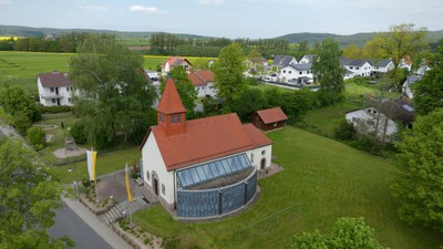 Ein weißes Gebäude mit rotem Dach in ländlicher Umgebung, umgeben von Grünflächen und weiteren Häusern.